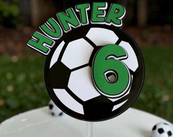 May include: A cake topper featuring a black and white soccer ball design with the name HUNTER in green letters arched above and a large green number 6 layered on the front. The topper is mounted on a clear stick and stands upright.