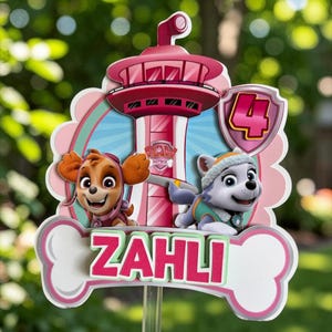 May include: A Paw Patrol themed cake topper featuring a pink lookout tower, the characters Skye and Everest, and a shield with the number 4. A white bone-shaped cutout at the bottom displays the name ZAHLI in bold pink letters.