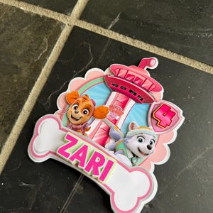 May include: A layered paper cake topper featuring Paw Patrol characters Skye and Everest, a pink lookout tower, and a large number 4. A white bone-shaped cutout at the bottom displays the name ZARI in bright pink letters with a green outline.