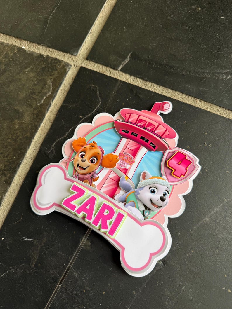 May include: A layered paper cake topper featuring Paw Patrol characters Skye and Everest, a pink lookout tower, and a large number 4. A white bone-shaped cutout at the bottom displays the name ZARI in bright pink letters with a green outline.