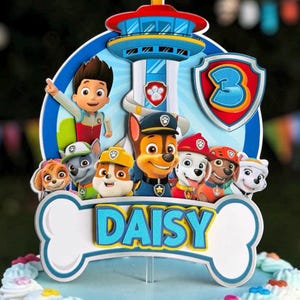 May include: A multi-layered cake topper featuring the Paw Patrol characters and the Lookout Tower. The design includes a blue shield with the number 3 and a white bone-shaped base with the name DAISY printed in blue and yellow letters.