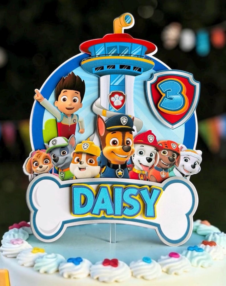 May include: A multi-layered cake topper featuring the Paw Patrol characters and the Lookout Tower. The design includes a blue shield with the number 3 and a white bone-shaped base with the name DAISY printed in blue and yellow letters.
