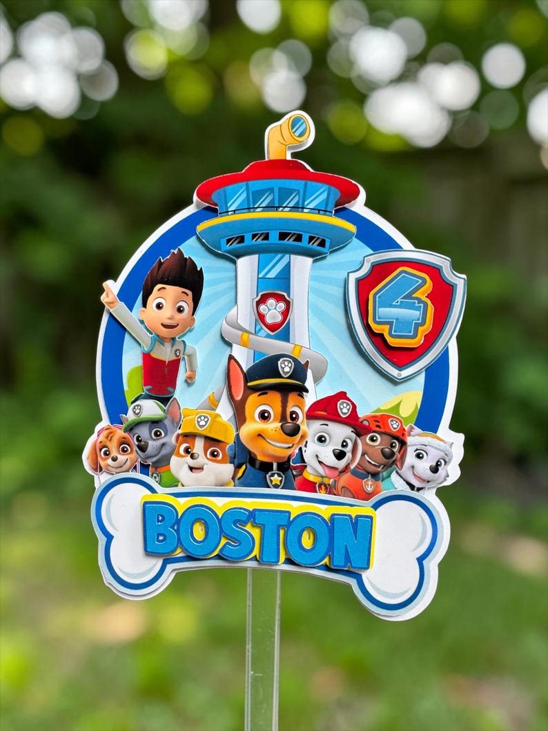 May include: A Paw Patrol themed cake topper featuring a central lookout tower, the character Ryder, and a group of puppies. The design includes a shield with the number 4 and a white bone-shaped base with the name BOSTON printed in blue letters.