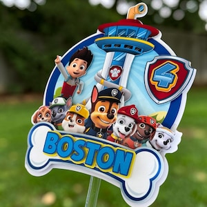 May include: A Paw Patrol themed cake topper featuring the Lookout tower, Ryder, and several pups. A shield with the number 4 is on the right. A white bone-shaped cutout at the bottom displays the name BOSTON in bold blue letters.