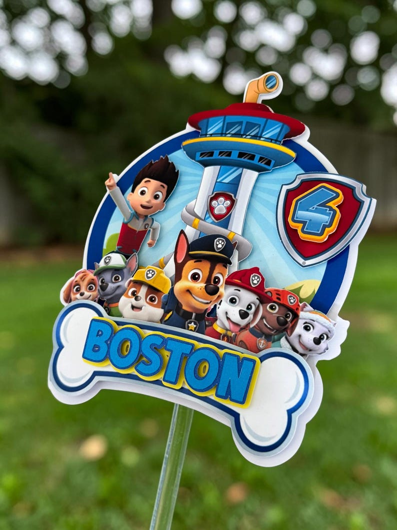 May include: A Paw Patrol themed cake topper featuring the Lookout tower, Ryder, and several pups. A shield with the number 4 is on the right. A white bone-shaped cutout at the bottom displays the name BOSTON in bold blue letters.