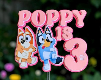 May include: A layered cake topper featuring the text "POPPY IS 3" in pink and white. The design includes two cartoon dog characters, one orange and white and one blue and white, positioned below the text. A small paw print is visible on the number 3.