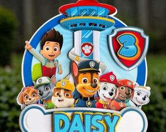 May include: A multi-layered cake topper featuring the Paw Patrol characters and the Lookout Tower. The design includes a blue shield with the number 3 and a white bone-shaped base with the name DAISY printed in blue and yellow letters.