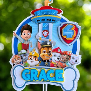May include: A Paw Patrol themed cake topper featuring a blue lookout tower, the character Ryder, and several puppies. A shield with the number 4 is on the right. A white bone-shaped banner at the bottom displays the name GRACE in blue letters.