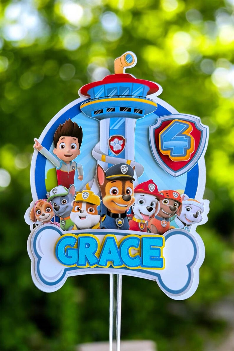 May include: A Paw Patrol themed cake topper featuring a blue lookout tower, the character Ryder, and several puppies. A shield with the number 4 is on the right. A white bone-shaped banner at the bottom displays the name GRACE in blue letters.