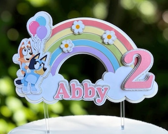 May include: A cake topper featuring a pastel rainbow, white clouds, and two cartoon dogs. The name Abby is written in pink letters with silver glitter accents. A large pink number 2 is on the right side, and three small white flowers are on the rainbow.