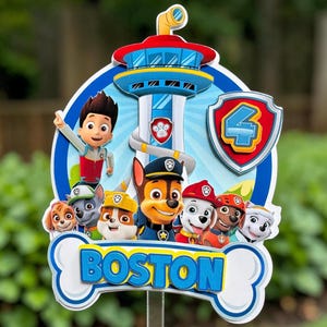May include: A multi-layered cake topper featuring the Paw Patrol characters and the Lookout tower. The design includes a shield with the number 4 and a bone-shaped banner at the bottom with the name BOSTON in blue 3D lettering.