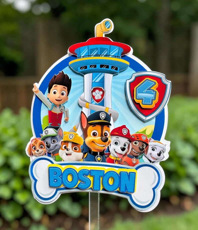 May include: A multi-layered cake topper featuring the Paw Patrol characters and the Lookout tower. The design includes a shield with the number 4 and a bone-shaped banner at the bottom with the name BOSTON in blue 3D lettering.