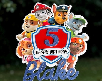 May include: A cake topper featuring Paw Patrol characters surrounding a red shield with a blue glittery number 5 and the text HAPPY BIRTHDAY. Below the shield, the name Blake is written in blue glittery script on a clear plastic stick.
