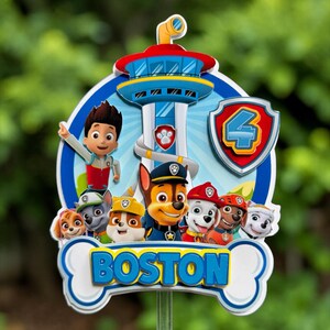May include: A Paw Patrol themed cake topper featuring Ryder and the puppy characters in front of the Lookout tower. The design includes a red shield with the number 4 and a white bone-shaped banner at the bottom with the name BOSTON in blue.