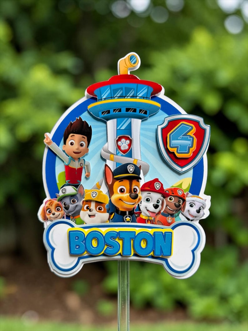 May include: A Paw Patrol themed cake topper featuring Ryder and the puppy characters in front of the Lookout tower. The design includes a red shield with the number 4 and a white bone-shaped banner at the bottom with the name BOSTON in blue.