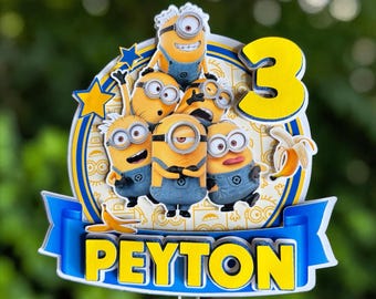 May include: A layered cake topper featuring five yellow Minion characters in blue overalls, a large yellow number 3, and a blue banner with the name PEYTON in yellow letters. The design includes yellow stars and banana illustrations on a white background.