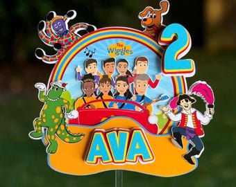 May include: A multi-layered cake topper featuring The Wiggles characters, a red car, a rainbow, and a brown dog. The topper displays the number 2 in blue and the name AVA in blue letters on an orange base, all mounted on a clear plastic stick.