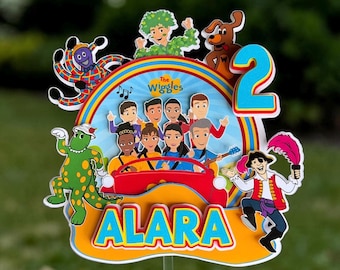 May include: A multi-layered cake topper featuring The Wiggles characters, a red car, a rainbow arch, and the name Alara in blue letters. The topper includes a large blue number 2, a green dinosaur, a brown dog, and a pirate figure.