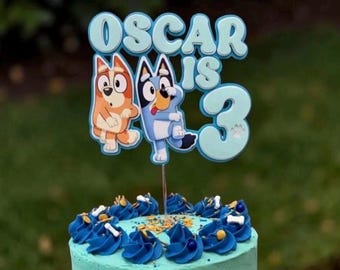May include: A light blue cake topper featuring the text OSCAR IS 3 with cartoon dog characters in orange and blue. The topper is positioned on a light blue frosted cake with dark blue piped frosting swirls and white bone decorations.