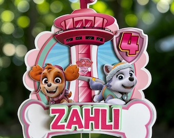 May include: A decorative cake topper featuring a pink Paw Patrol lookout tower, the characters Skye and Everest, a shield with the number 4, and a white bone-shaped banner with the name ZAHLI printed in pink letters on a light green background.