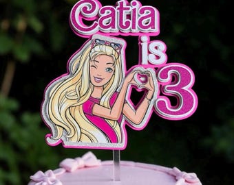 May include: A cake topper featuring a cartoon character with long blonde hair, wearing a pink shirt and sunglasses on her head, making a heart shape with her hands. The text reads Catia is 3 in glittery pink and silver layered lettering.