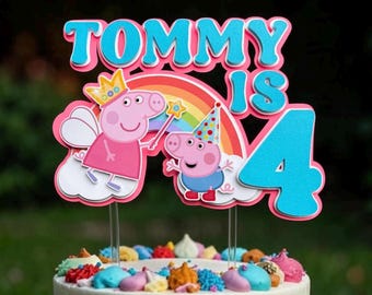 May include: A cake topper featuring the text TOMMY IS 4 in blue and pink. It displays two pink pig characters, one wearing a yellow crown and holding a star wand, the other wearing a colorful party hat. A rainbow and white clouds are behind them.
