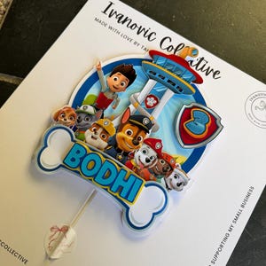 May include: A custom cake topper featuring Paw Patrol characters and the name BODHI in blue letters on a white bone-shaped cutout. The design includes a blue shield with the number 3 and a lookout tower, all mounted on a thin white stick.