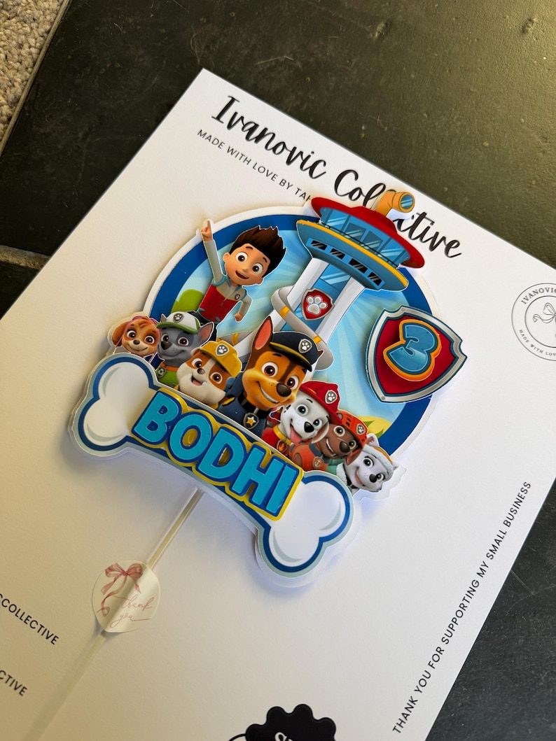 May include: A custom cake topper featuring Paw Patrol characters and the name BODHI in blue letters on a white bone-shaped cutout. The design includes a blue shield with the number 3 and a lookout tower, all mounted on a thin white stick.