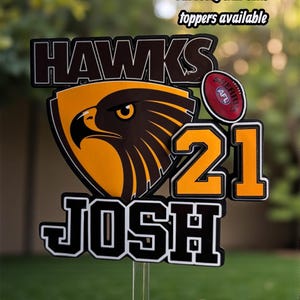 May include: A custom cake topper featuring the word HAWKS in black, a hawk emblem, the number 21, and the name JOSH in black and white. A small red Sherrin AFL football is attached. Text at the top reads All footy club cake toppers available.