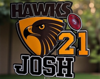 May include: A cake topper featuring the text HAWKS, JOSH, and the number 21, with a hawk emblem and a small red Sherrin AFL football. The text All footy club cake toppers available is printed at the top of the design in white letters.