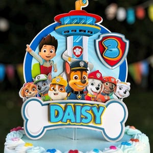 May include: A multi-layered cake topper featuring Paw Patrol characters including Ryder, Chase, Marshall, Rubble, Skye, Rocky, and Zuma. The design includes a lookout tower, a shield with the number 3, and a bone-shaped sign reading DAISY.