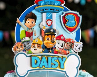 May include: A multi-layered cake topper featuring Paw Patrol characters including Ryder, Chase, Marshall, Rubble, Skye, Rocky, and Zuma. The design includes a lookout tower, a shield with the number 3, and a bone-shaped sign reading DAISY.