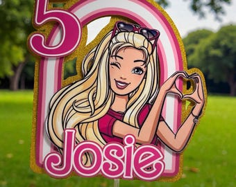 May include: A glittery gold cake topper featuring a cartoon character with blonde hair, sunglasses, and a pink shirt. The design includes a large pink number 5, a rainbow arch, and the name Josie in pink text. The character is making a heart shape.