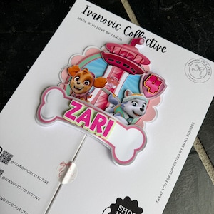 May include: A custom cake topper featuring Paw Patrol characters Skye and Everest, a pink lookout tower, and a large number 4. A white bone-shaped cutout displays the name ZARI in bright pink letters. The topper is mounted on a thin stick.