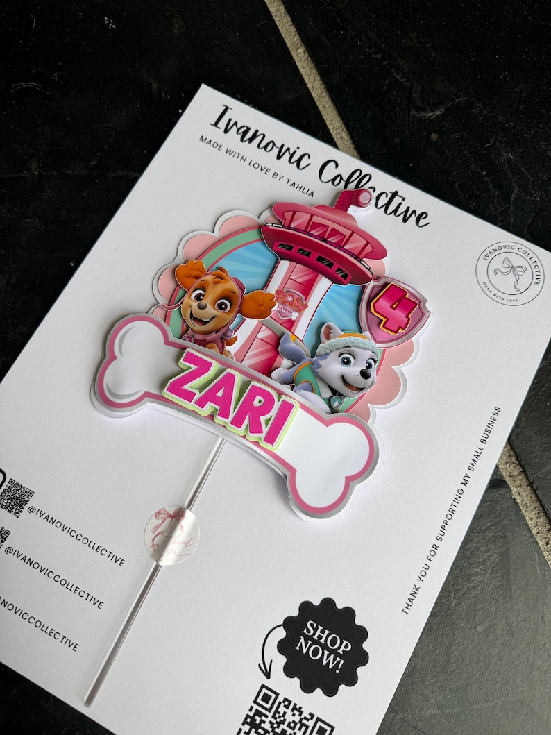 May include: A custom cake topper featuring Paw Patrol characters Skye and Everest, a pink lookout tower, and a large number 4. A white bone-shaped cutout displays the name ZARI in bright pink letters. The topper is mounted on a thin stick.