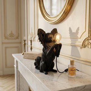 Può includere: Una lampada da tavolo nero opaco a forma di cane Papillon seduto con grandi orecchie piumate. La figura del cane tiene una lampadina in vetro trasparente tra le zampe anteriori, collegata tramite un cavo nero con interruttore a una presa.