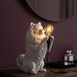 Op de afbeelding: Een wit, sculpturaal Pomeranian-hondje dat dienst doet als tafellamp. De hond zit rechtop en houdt tussen zijn voorpoten een zwarte fitting vast met een gloeiende, warmkleurige Edison-gloeilamp. Een zwart netsnoer loopt vanaf de basis weg.