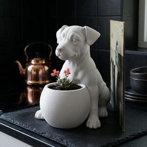 Può includere: Un vaso in ceramica bianca opaca a forma di cane seduto, dotato di una base rotonda a forma di ciotola che contiene una piccola pianta succulenta verde con foglie dalle punte arancioni. La figura del cane presenta tratti del viso dettagliati.