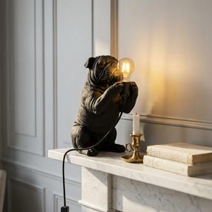 Op de afbeelding: Een donker antracietgrijze bulldog-figuurlamp die een gloeiende ronde Edison-stijl gloeilamp in zijn voorpoten houdt. De lamp heeft een zwart netsnoer met een schakelaar en staat naast een koperen kandelaar en twee op elkaar gestapelde boeken.