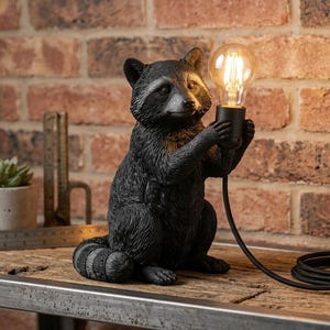 Op de afbeelding: Een zwart-grijze tafellamp in de vorm van een wasbeer met een textuur van vacht. De wasbeerfiguur houdt een zwarte fitting vast met een gloeiende Edison-lamp. Een zwart netsnoer met een schakelaar loopt vanaf de basis van de lamp.