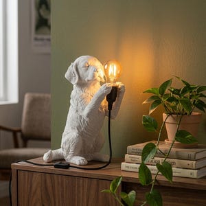 May include: A white dog-shaped table lamp featuring a textured finish, sitting in a seated position while holding a black socket with a glowing, warm-toned Edison-style light bulb. The lamp includes a black power cord with an inline switch.