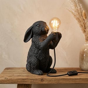 Könnte beinhalten: Eine schwarze Tischleuchte in Hasenform mit strukturierter Felloptik. Der Hase sitzt und hält eine klare Glühbirne in seinen Vorderpfoten. Ein schwarzes Stromkabel mit einem Zwischenschalter führt von der Lampenfassung weg.