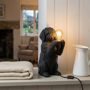 Op de afbeelding: Een zwarte tafellamp in de vorm van een hond met een getextureerde vacht, rechtop zittend en een heldere, gloeiende Edison-stijl gloeilamp tussen de voorpoten houdend. De lamp heeft een zwart snoer met een schakelaar aan de basis.