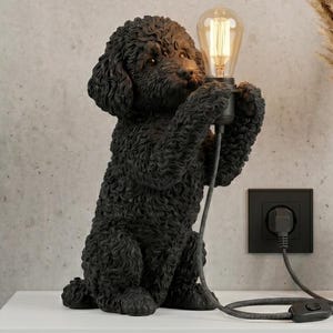 May include: A black dog-shaped table lamp with a textured, curly-haired finish. The dog is sitting upright and holding a clear, vintage-style Edison light bulb in its front paws. A black power cord with an inline switch extends from the bulb base.