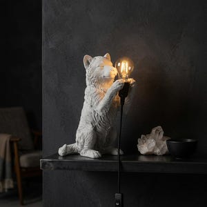May include: A white, textured wolf-shaped table lamp standing on its hind legs. The wolf holds a black socket with a glowing, clear glass Edison-style light bulb. A black power cord with an inline switch hangs down from the lamp's base.