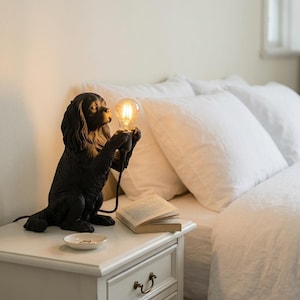 May include: A black dog-shaped table lamp with a textured finish, depicted in a sitting position while holding a glowing, clear glass Edison-style light bulb between its front paws. The lamp features detailed fur sculpting and a black power cord.
