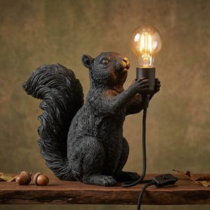 May include: A black squirrel-shaped table lamp featuring a textured fur finish. The squirrel is standing upright and holding a clear glass Edison-style light bulb in its front paws. A black power cord with an inline switch extends from the bulb base.