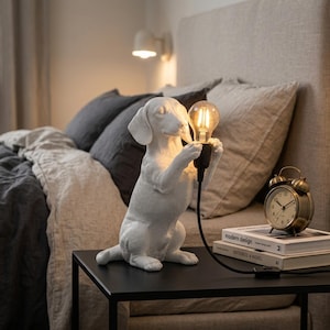 Op de afbeelding: Een witte, matte tafellamp in de vorm van een teckel die op zijn achterpoten staat en een zwarte fitting met een brandende Edison-gloeilamp vasthoudt. De lamp heeft een zwart snoer met een schakelaar en staat op een zwarte bijzettafel.