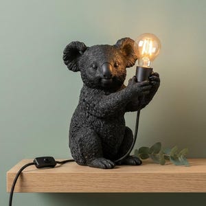 Può includere: Una lampada da tavolo a forma di koala nero con finitura testurizzata. La figura del koala è seduta in posizione eretta e regge un portalampada nero con una lampadina rotonda in vetro trasparente accesa. Un cavo nero con interruttore.
