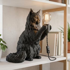 Könnte beinhalten: Eine schwarze Tischleuchte in Katzenform mit strukturierter Fellzeichnung. Die Katze sitzt aufrecht und hält eine dunkelgraue Fassung mit einer klaren Edison-Glühbirne. Ein schwarzes, gedrehtes Stoffkabel mit Schalter führt nach unten.